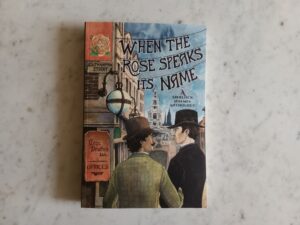 Front cover of When the Rose Speaks Its Name. When the Rose Speaks Its Name, in lettering hanging from telegraph wires. The streets of London with signs reading Southampton Street and Geo: Newnes Ltd (limited) Offices with a drawn hand pointing to the left. Watson and Holmes are in the foreground. Watson is wearing a light green check jacket and a brown bowler. Holmes is wearing a deep black jacket with purple cravat showing and a tall black homburg. Holmes and Watson have their arms linked and are looking into each others eyes.