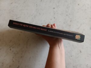 Hand holding paperback edition of When the Rose Speaks Its Name, showing the binding